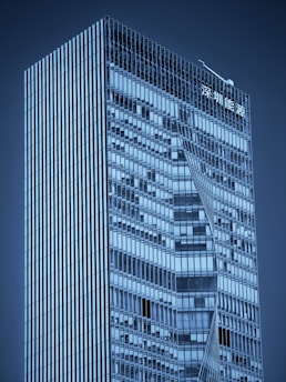 A modern skyscraper with a sleek, glass exterior featuring geometric patterns. The building has a high-rise structure with sharp lines and reflective windows. There is signage in an Asian script at the top.