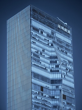 A modern skyscraper with a sleek, glass exterior featuring geometric patterns. The building has a high-rise structure with sharp lines and reflective windows. There is signage in an Asian script at the top.