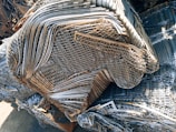 A large pile of intricately layered metal grids and sheets, likely indicating a scrap or recycling yard. The metal consists of various shades of gray, silver, and rusted brown, showing signs of weathering and age.