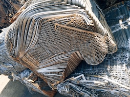 A large pile of intricately layered metal grids and sheets, likely indicating a scrap or recycling yard. The metal consists of various shades of gray, silver, and rusted brown, showing signs of weathering and age.