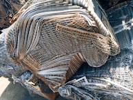 A large pile of intricately layered metal grids and sheets, likely indicating a scrap or recycling yard. The metal consists of various shades of gray, silver, and rusted brown, showing signs of weathering and age.