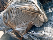 A large pile of intricately layered metal grids and sheets, likely indicating a scrap or recycling yard. The metal consists of various shades of gray, silver, and rusted brown, showing signs of weathering and age.