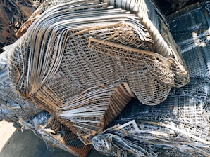 A large pile of intricately layered metal grids and sheets, likely indicating a scrap or recycling yard. The metal consists of various shades of gray, silver, and rusted brown, showing signs of weathering and age.