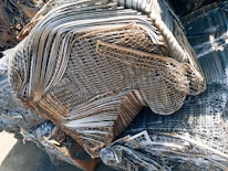 A large pile of intricately layered metal grids and sheets, likely indicating a scrap or recycling yard. The metal consists of various shades of gray, silver, and rusted brown, showing signs of weathering and age.