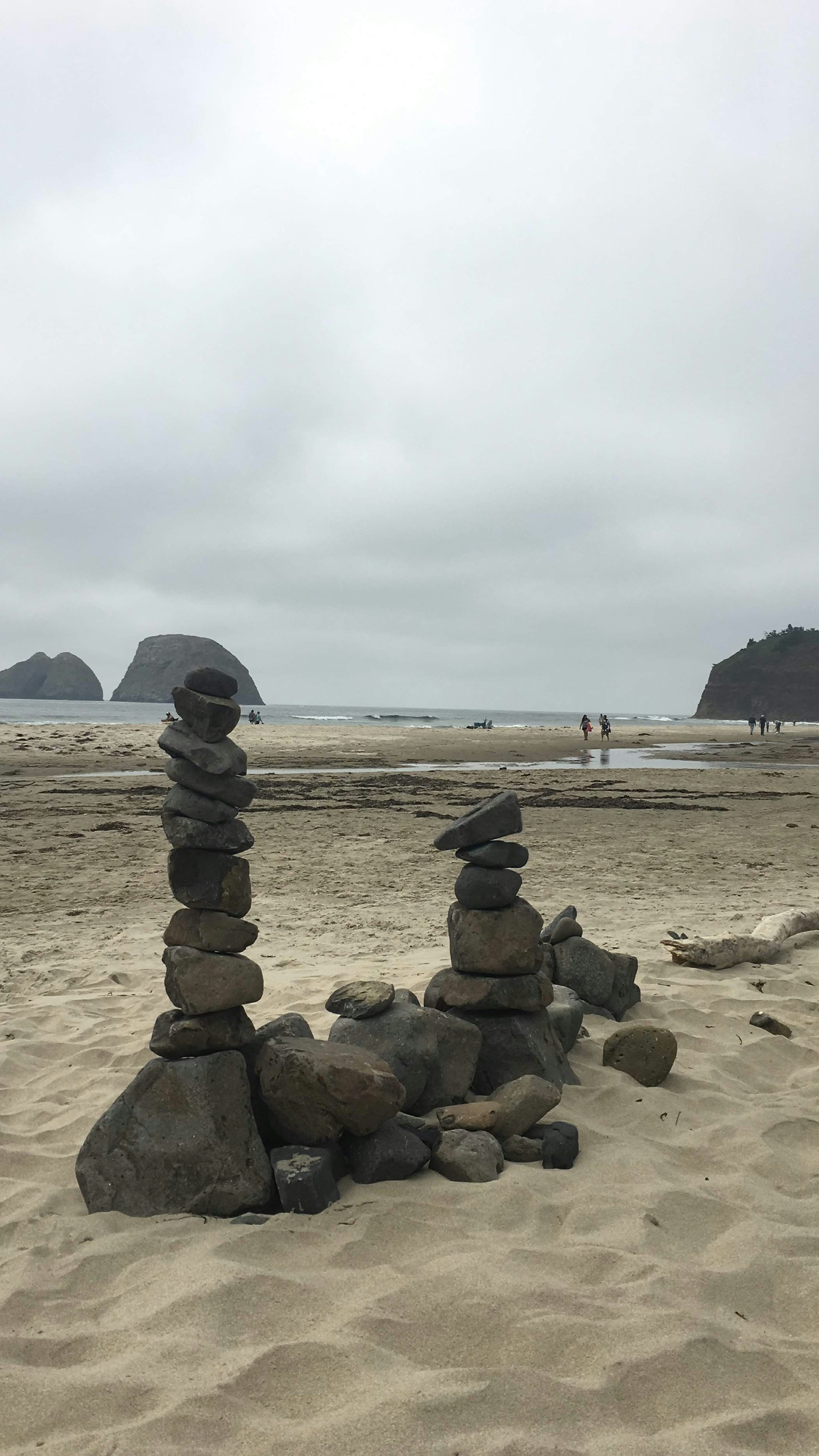 A beach scene with stacks of stones arranged artfully in the foreground. The sandy shore extends to the horizon where the ocean meets a cloudy sky. In the background, there are distant islands and a few people walking along the beach.