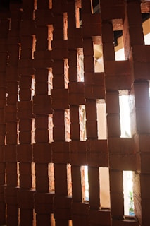 A textured wall made of interlocking reddish-brown bricks with openings allowing light to pass through. The bricks are arranged in a geometric pattern, creating a play of light and shadows.