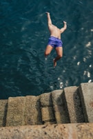 A person is diving mid-air towards the water from a set of stone steps. The individual is wearing blue swim shorts. The water appears dark and subtly reflective, while the stone steps provide a textured, earthy contrast.
