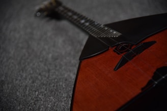 A close-up of a string instrument laying on a textured gray fabric surface. The instrument features a wooden, reddish-brown body with a black triangular section at the top. Strings and tuning pegs are visible, extending from the body toward the blurred neck in the background.