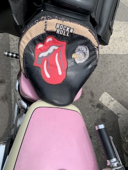 A motorcycle seat cover features a prominent red and white logo of a tongue and lips, the phrase 'ROCK N ROLL,' and an illustration of an eagle wearing sunglasses. The seat sits atop a pink motorcycle with chrome handlebars, parked on an asphalt surface.
