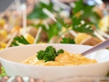 A creamy yellow dip is served in a white bowl, garnished with fresh parsley. The focus is on the dip, which has a smooth texture. In the background, there are skewers with visible bits of food and hints of greenery, creating a fresh and inviting atmosphere.