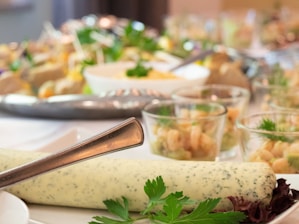 A close-up of a table filled with a variety of food items, including a large log of herb-infused butter in the foreground, surrounded by parsley leaves. In the background, there are several small glass bowls filled with different dishes, possibly salads or appetizers, garnished with herbs. The setting appears to be elegant and catered.