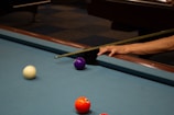 A close-up view of a pool table corner where a person's hand is holding a cue stick. A white ball is moving towards the purple ball near the pocket, and a vibrant orange-red ball is near the bottom of the frame. The surface of the pool table is blue, and a dark brown wooden edge is visible.