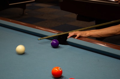 A close-up view of a pool table corner where a person's hand is holding a cue stick. A white ball is moving towards the purple ball near the pocket, and a vibrant orange-red ball is near the bottom of the frame. The surface of the pool table is blue, and a dark brown wooden edge is visible.