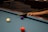 A close-up view of a pool table corner where a person's hand is holding a cue stick. A white ball is moving towards the purple ball near the pocket, and a vibrant orange-red ball is near the bottom of the frame. The surface of the pool table is blue, and a dark brown wooden edge is visible.
