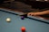 A close-up view of a pool table corner where a person's hand is holding a cue stick. A white ball is moving towards the purple ball near the pocket, and a vibrant orange-red ball is near the bottom of the frame. The surface of the pool table is blue, and a dark brown wooden edge is visible.