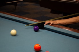 A close-up view of a pool table corner where a person's hand is holding a cue stick. A white ball is moving towards the purple ball near the pocket, and a vibrant orange-red ball is near the bottom of the frame. The surface of the pool table is blue, and a dark brown wooden edge is visible.