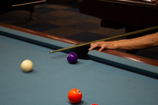 A close-up view of a pool table corner where a person's hand is holding a cue stick. A white ball is moving towards the purple ball near the pocket, and a vibrant orange-red ball is near the bottom of the frame. The surface of the pool table is blue, and a dark brown wooden edge is visible.