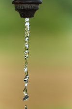 A close-up of water flowing from an outdoor faucet, captured in a still moment with a clear background. The water stream is transparent, exhibiting ripples and reflections.