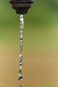 A close-up of water flowing from an outdoor faucet, captured in a still moment with a clear background. The water stream is transparent, exhibiting ripples and reflections.