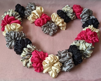 A heart-shaped arrangement of satin scrunchies in various colors including pink, cream, black, and gray, placed on a textured pink fabric background. The scrunchies are ruched and layered, creating a fluffy and voluminous effect.