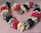 A heart-shaped arrangement of satin scrunchies in various colors including pink, cream, black, and gray, placed on a textured pink fabric background. The scrunchies are ruched and layered, creating a fluffy and voluminous effect.