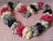 A heart-shaped arrangement of satin scrunchies in various colors including pink, cream, black, and gray, placed on a textured pink fabric background. The scrunchies are ruched and layered, creating a fluffy and voluminous effect.
