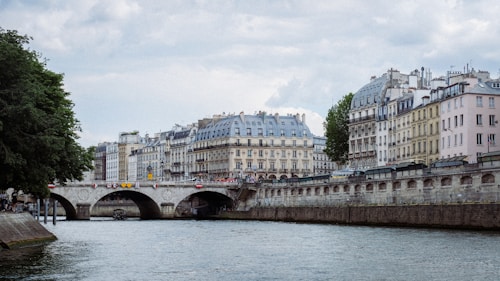 A serene European urban scene featuring a river with a stone bridge arching over it, lined by classic multi-story buildings with French architecture along the riverside. The sky is overcast, and there are green trees visible to the left, adding a touch of nature to the scene.
