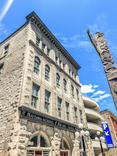 A historic stone building with arched windows houses the Ottawa School of Art. A totem pole stands adjacent, and a parking garage spirals upward in the background. The sky is bright with a few clouds.
