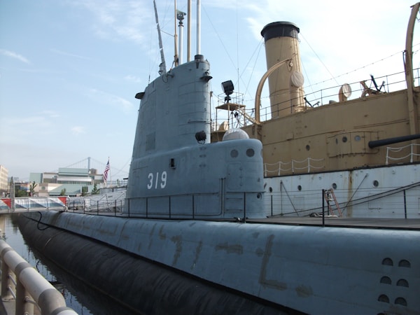A grey submarine is docked next to an older, larger ship. The submarine has the number 319 painted on its side. In the background, there are modern buildings and an American flag is visible, indicating a dock or museum setting.