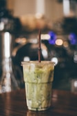 A plastic cup filled with a green matcha latte is placed on a wooden surface. The cup has a brown straw and the drink appears to be iced, as indicated by the visible ice cubes. The background is blurred with warm, ambient lighting, creating a cozy atmosphere.