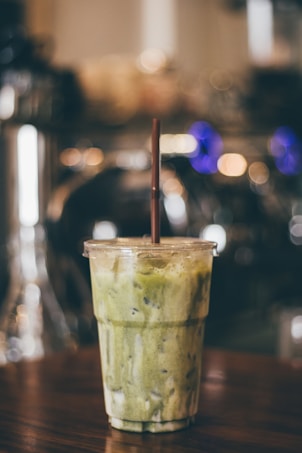 A plastic cup filled with a green matcha latte is placed on a wooden surface. The cup has a brown straw and the drink appears to be iced, as indicated by the visible ice cubes. The background is blurred with warm, ambient lighting, creating a cozy atmosphere.
