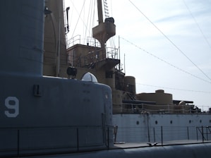 A detailed view of a naval vessel, featuring large metal structures and various deck equipment. The ship has a number 9 marked on its side, and there are ropes and other nautical elements visible. The lighting suggests a calm, possibly overcast day.