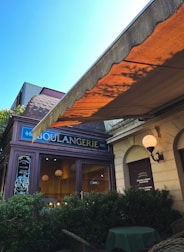 A charming storefront with a traditional awning and a sign reading 'Boulangerie' in an elegant font. The facade features a mix of classical and rustic architecture, with a hanging street lamp and decorative plants in front. Inside, warm lighting and spherical lamps create a cozy atmosphere.