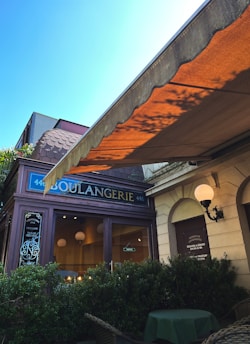 A charming storefront with a traditional awning and a sign reading 'Boulangerie' in an elegant font. The facade features a mix of classical and rustic architecture, with a hanging street lamp and decorative plants in front. Inside, warm lighting and spherical lamps create a cozy atmosphere.