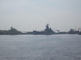 A large battleship is docked at a harbor, surrounded by cranes and industrial structures. The sky is overcast, and the water appears calm with gentle ripples extending across the surface. In the background, trees can be seen on the left, and several other ships are visible docked along the harbor.