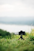 A camera is positioned on a small tripod in a grassy outdoor setting with hills and sky in the background, giving a sense of open space and nature.