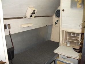 A small, enclosed room featuring a bunk bed with a grey mattress on one side. Above the bed, there is a control panel with dials and switches. Adjacent to the bed, there's a compact workspace with a small shelf and a desk. A photo frame and cap are placed on the desk. The walls are light-colored, giving the space a minimalist and utilitarian feel.