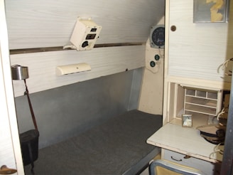 A small, enclosed room featuring a bunk bed with a grey mattress on one side. Above the bed, there is a control panel with dials and switches. Adjacent to the bed, there's a compact workspace with a small shelf and a desk. A photo frame and cap are placed on the desk. The walls are light-colored, giving the space a minimalist and utilitarian feel.