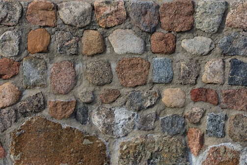 A wall constructed from an assortment of irregularly shaped stones in various earthy tones, including brown, gray, and beige. The stones are held together with mortar, creating a rustic and textured appearance.