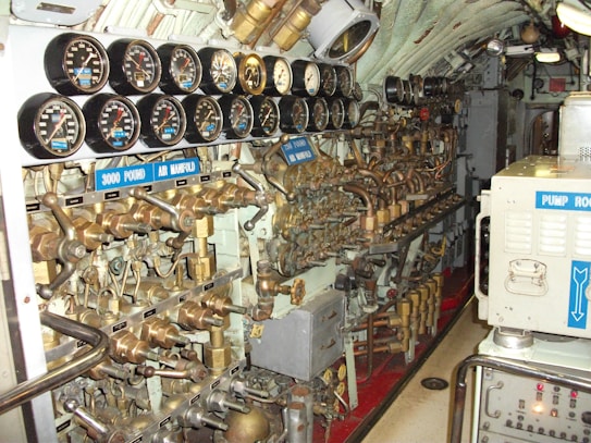 A complex array of gauges, valves, and pipes fills the submarine's control room. The dials display various pressure readings, and the intricate arrangement of brass machinery suggests a highly technical environment. Labels such as '3000 Pound Air Manifold' and 'Pump Room' indicate the functionality related to air and pumps.