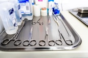 A stainless steel tray is arranged with various surgical instruments and medical supplies. There are pairs of scissors, forceps, and a pair of tweezers. In the background, there are bottles of antiseptic solution and medical tape.