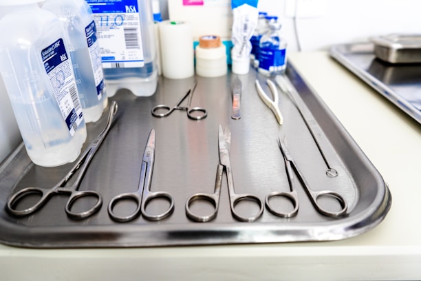 A stainless steel tray is arranged with various surgical instruments and medical supplies. There are pairs of scissors, forceps, and a pair of tweezers. In the background, there are bottles of antiseptic solution and medical tape.