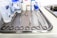A stainless steel tray is arranged with various surgical instruments and medical supplies. There are pairs of scissors, forceps, and a pair of tweezers. In the background, there are bottles of antiseptic solution and medical tape.
