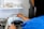 A person is operating a mini high temperature sterilizer in a clinical or laboratory setting. The person is holding a pair of scissors near the sterilizer, which is placed on a metal surface. Shelves above hold various supplies, including cotton swabs and small containers.