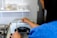 A person is operating a mini high temperature sterilizer in a clinical or laboratory setting. The person is holding a pair of scissors near the sterilizer, which is placed on a metal surface. Shelves above hold various supplies, including cotton swabs and small containers.