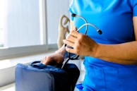 A person dressed in blue medical scrubs is holding a stethoscope in one hand and is placing or retrieving it from a medical bag with the other hand. The scene appears to be indoors near a window with a beige-colored object visible in the background.