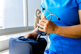 A person dressed in blue medical scrubs is holding a stethoscope in one hand and is placing or retrieving it from a medical bag with the other hand. The scene appears to be indoors near a window with a beige-colored object visible in the background.
