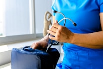 A person dressed in blue medical scrubs is holding a stethoscope in one hand and is placing or retrieving it from a medical bag with the other hand. The scene appears to be indoors near a window with a beige-colored object visible in the background.