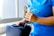 A person dressed in blue medical scrubs is holding a stethoscope in one hand and is placing or retrieving it from a medical bag with the other hand. The scene appears to be indoors near a window with a beige-colored object visible in the background.