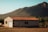 A solitary house with a reddish-brown tiled roof stands in a serene rural setting. It is surrounded by sparse vegetation and open land, with a backdrop of green, forested hills under a clear blue sky.